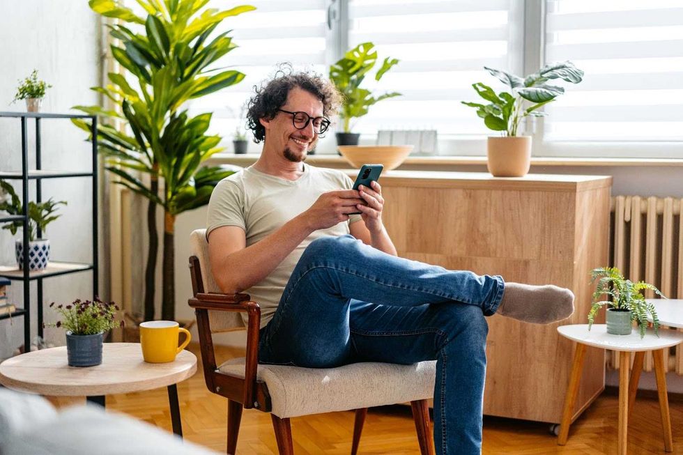 A man sitting in a brightly lit, modern space, smiles while looking at a phone.