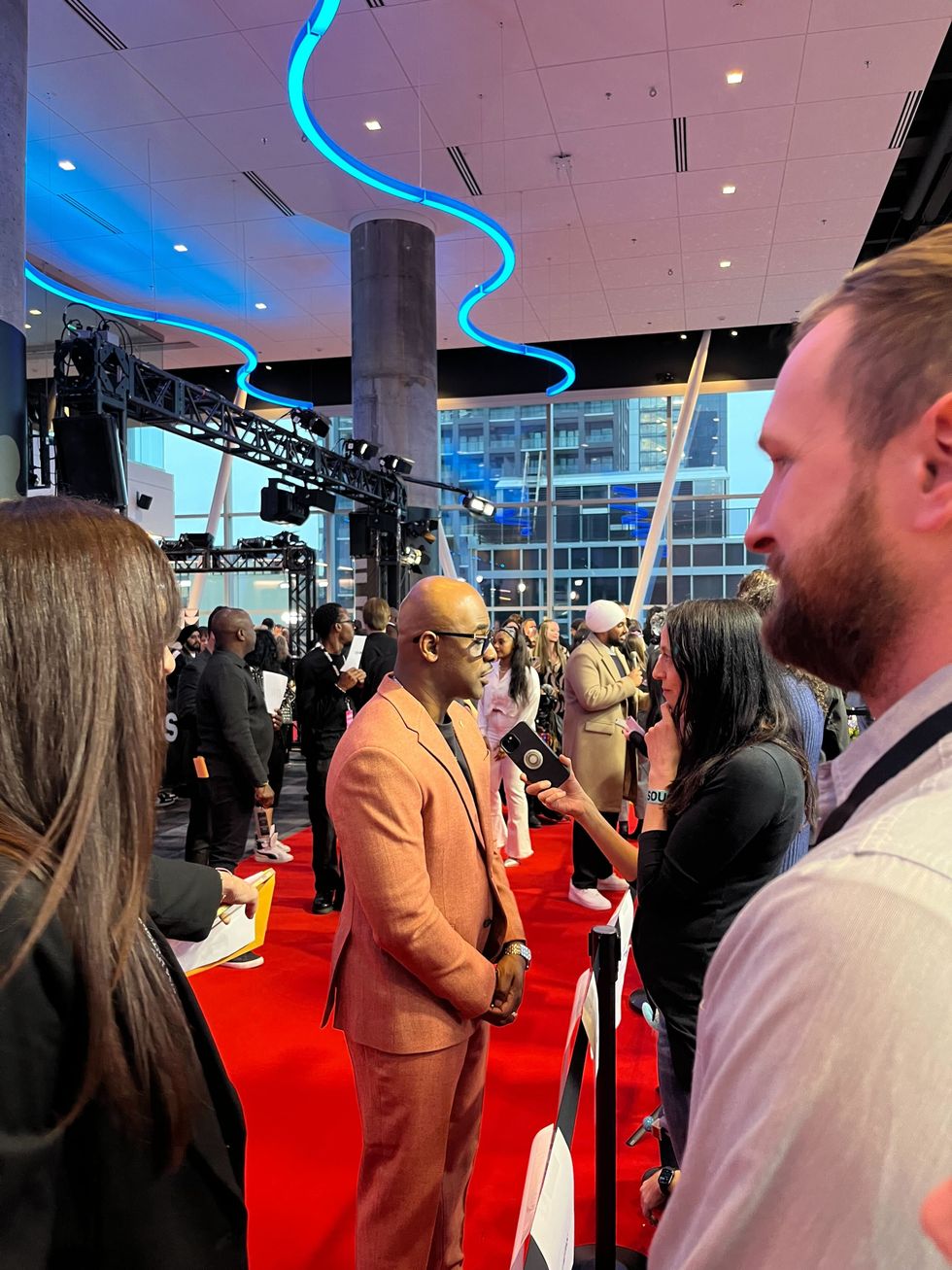 A man wearing a burnt orange suit speaking into a phone with people all around him on the red carpet.