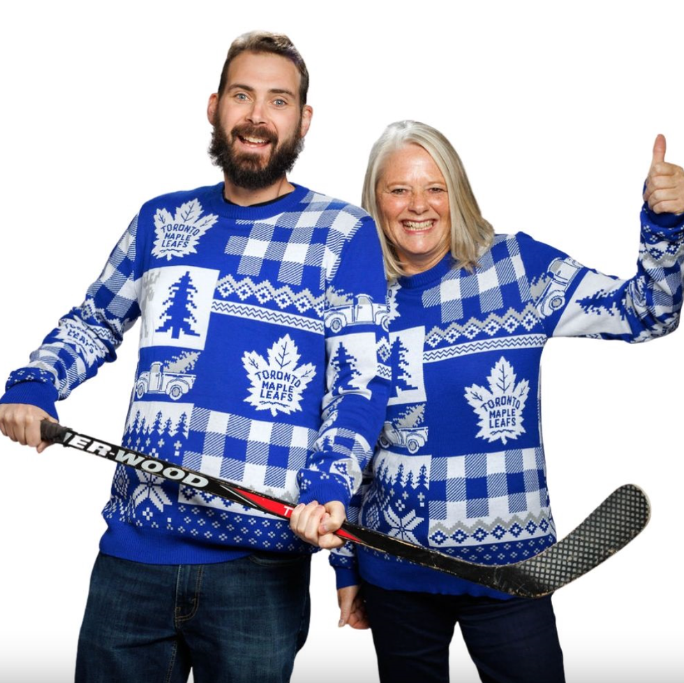 A man wearing a Toronto Maple Leafs ugly Christmas sweater holding a hockey stick and a woman standing next to him in the same sweater with a thumbs up.