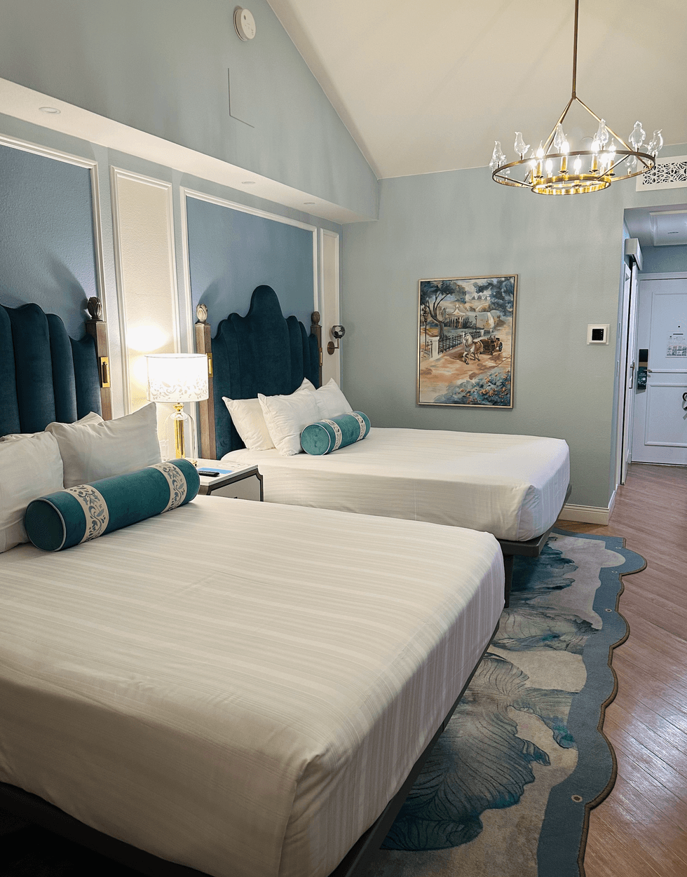 A Mary Poppins-themed standard room at Disney's Grand Floridian Resort, featuring two beds with teal velvet headboards, crisp white linens, teal bolster pillows, a gold chandelier with bird details, and a watercolour painting on the wall.