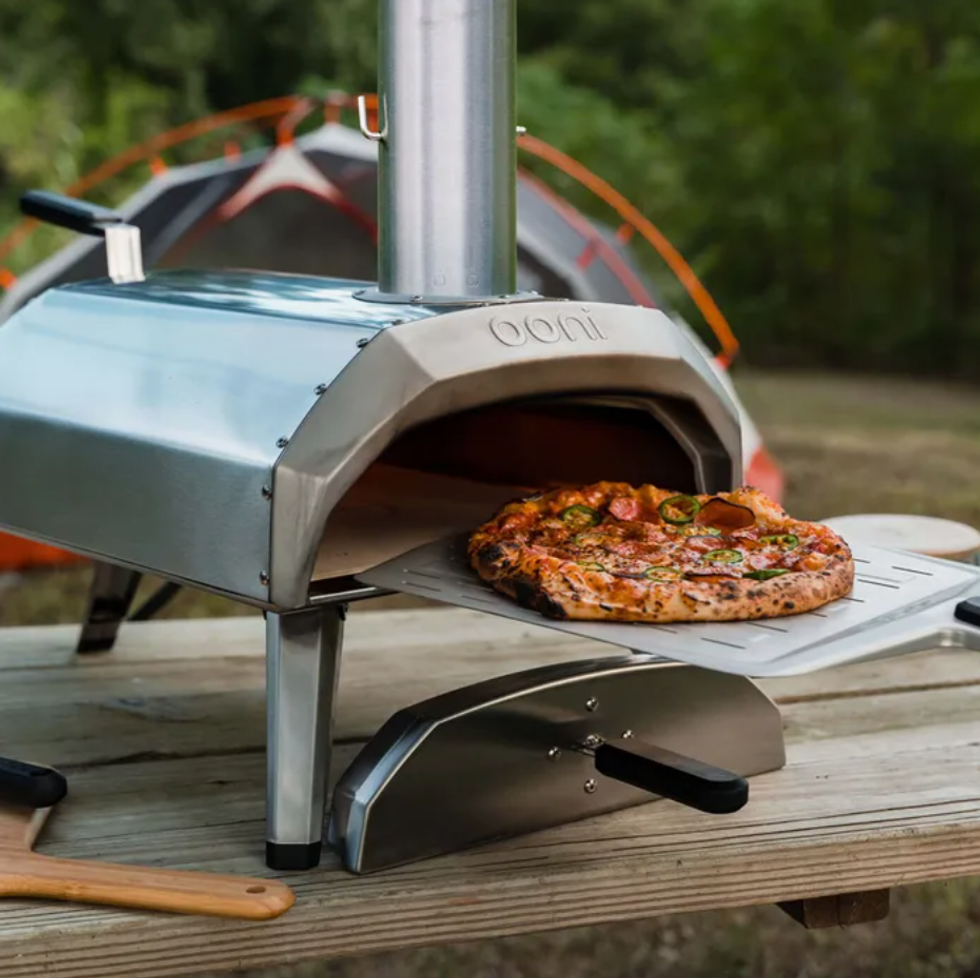 A metal camping pizza oven on a wooden picnic table with a cooked pizza on a steel pizza peel.