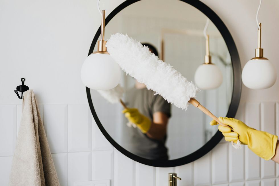 A mirror being dusted by a white feather duster held by someone wearing a yellow glove who is obscure in the mirror reflection.