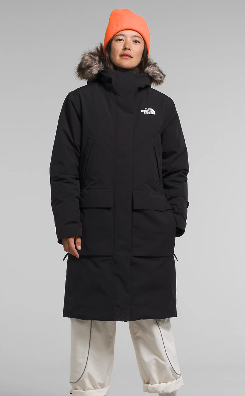 A model wearing a black winter jacket, white pants and orange hat.