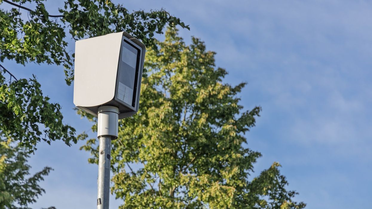A modern speed camera stands tall amidst green trees against a bright blue sky in the Netherlands.
