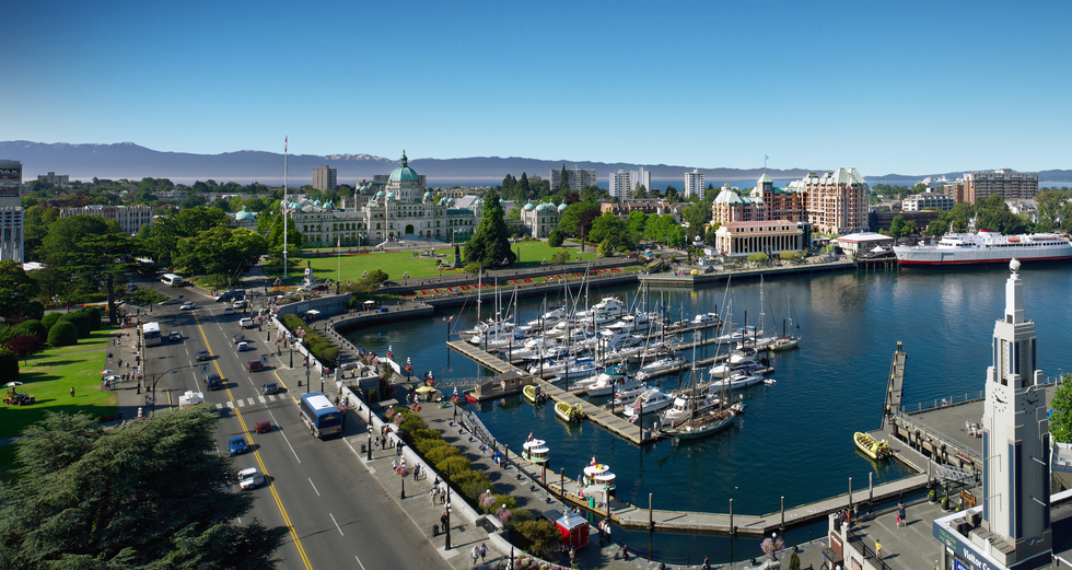 A panoramic photo of Victoria, BC with boats in the inner harbour and cars on the road and mountains in the background.
