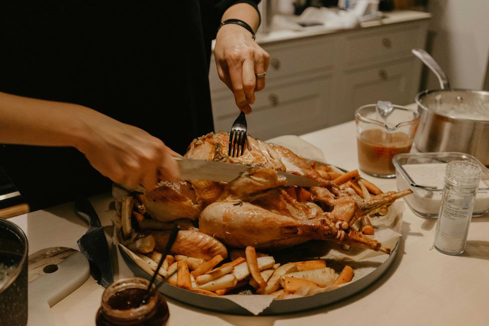 A person carves a turkey that sitting on a platter with roast vegetables, surrounded by seasonings, gravy and dressing.