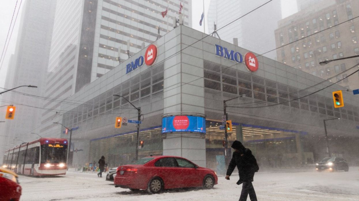 A person crossing the street with cars on the road and a building that says BMO on it in a snowstorm.