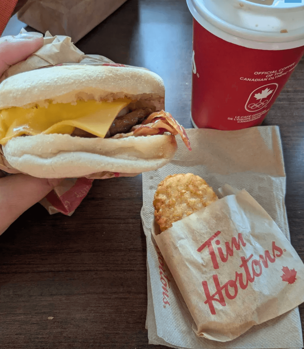 A person holding a breakfast sandwich and a hash brown on a table.