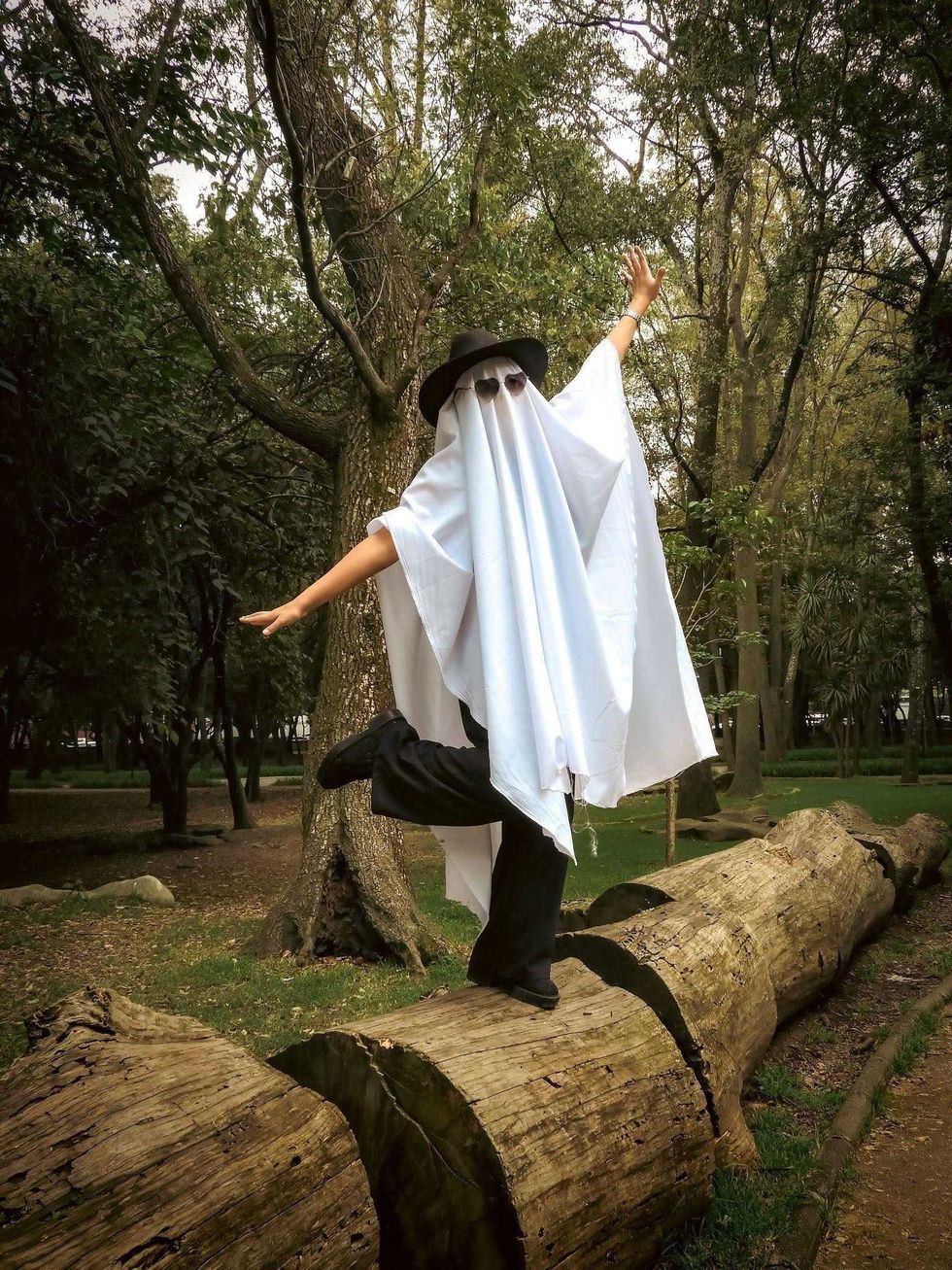 A person in a ghost costume, sunglasses and hat poses on a log.