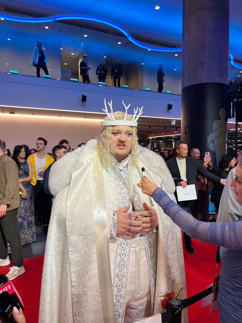 A person in an all-white outfit with a cape and antlers doing an interview at an event.