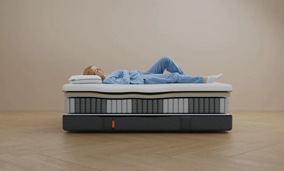 A person in blue pyjamas lays on top of an Emma Sleep Hybrid Comfort mattress. The view is from the side, and the layers of the mattress are illustrated to show how they offer support to the sleeping person.
