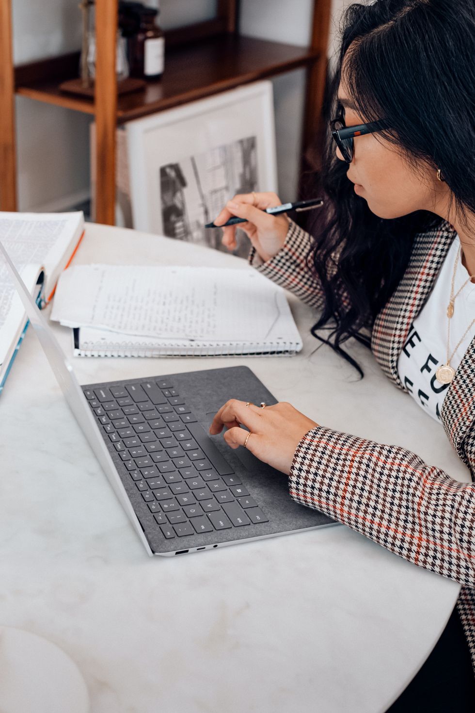 A person sitting at a dining table looking at their notes, holding a pen in one hand and controlling a laptop mouse with the other.