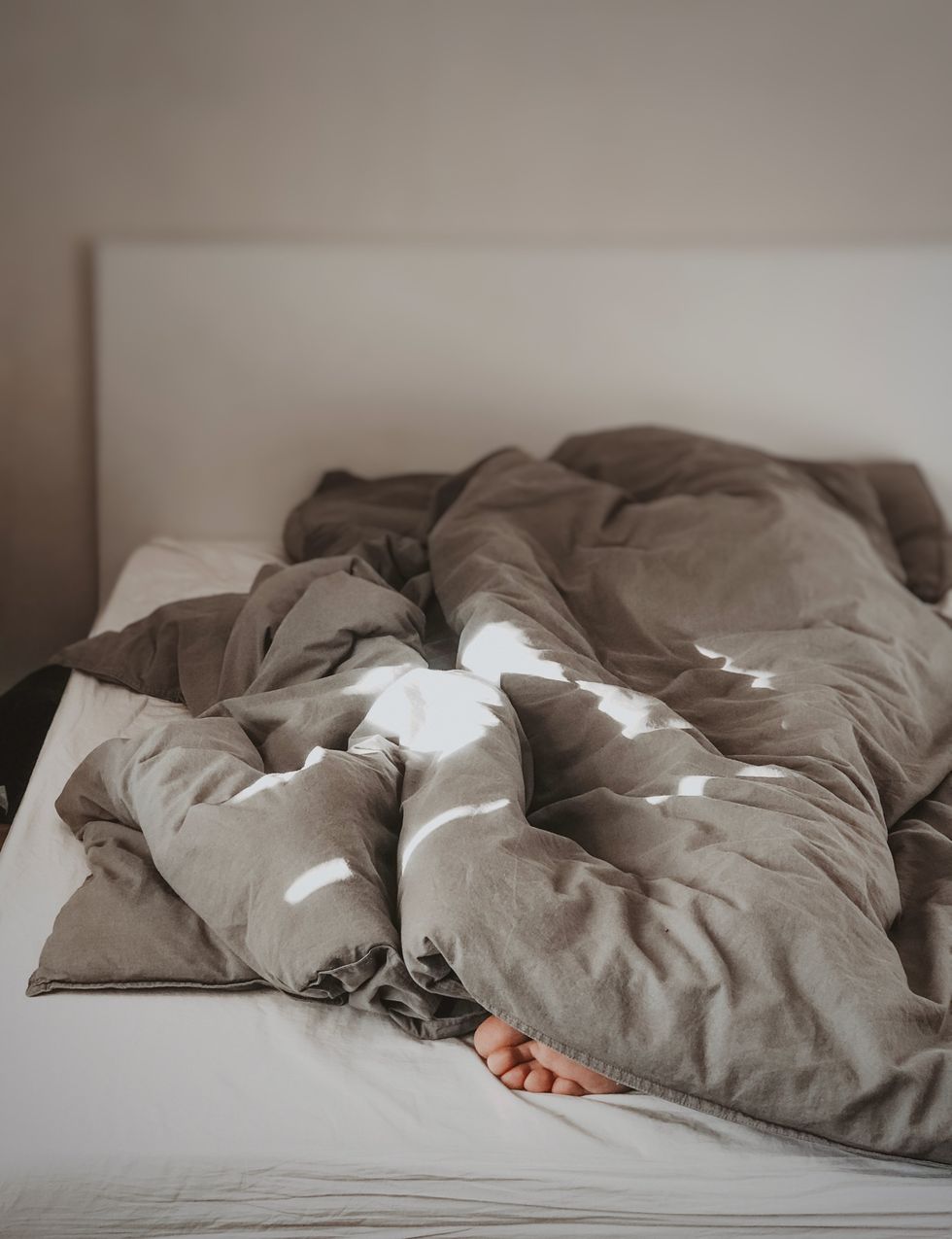A person sleeps underneath a grey comforter on the bed. All we can see of the person are their toes poking from under the comforter.