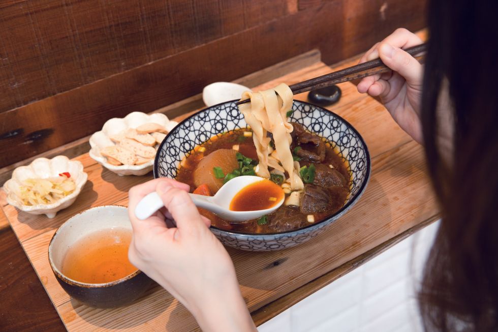 A person uses chopsticks and a spoon to eat noodle soup. 