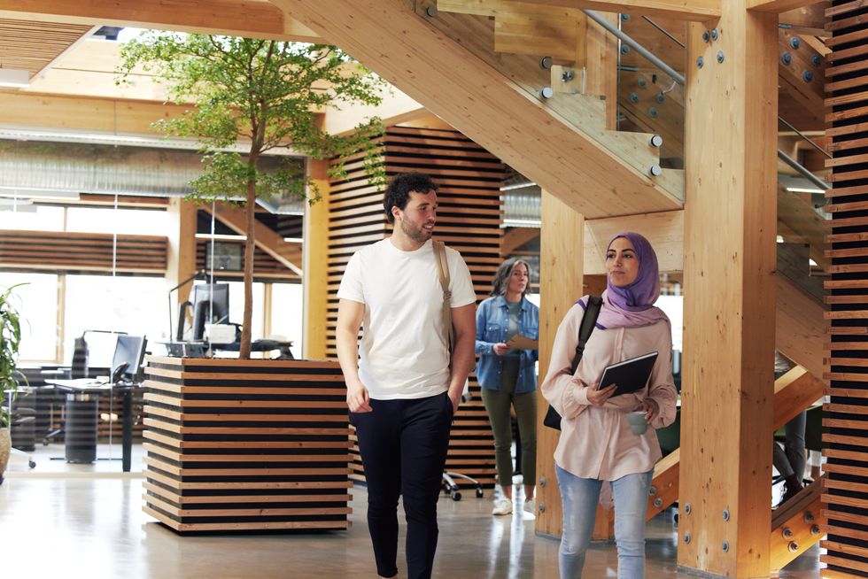 A person wearing a purple head scarf walks with another person through a corporate foyer, a person in a denim jacket stands in the background.