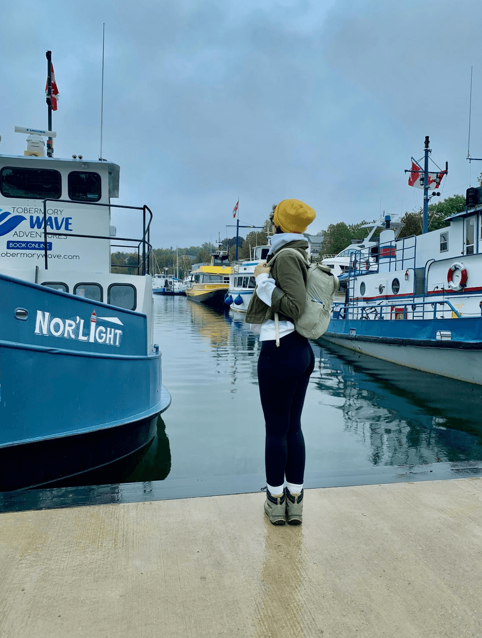 A person wearing hiking boots and a yellow hat standing beside boats in Tobermory's Little Tub Harbour on a cloudy fall day.