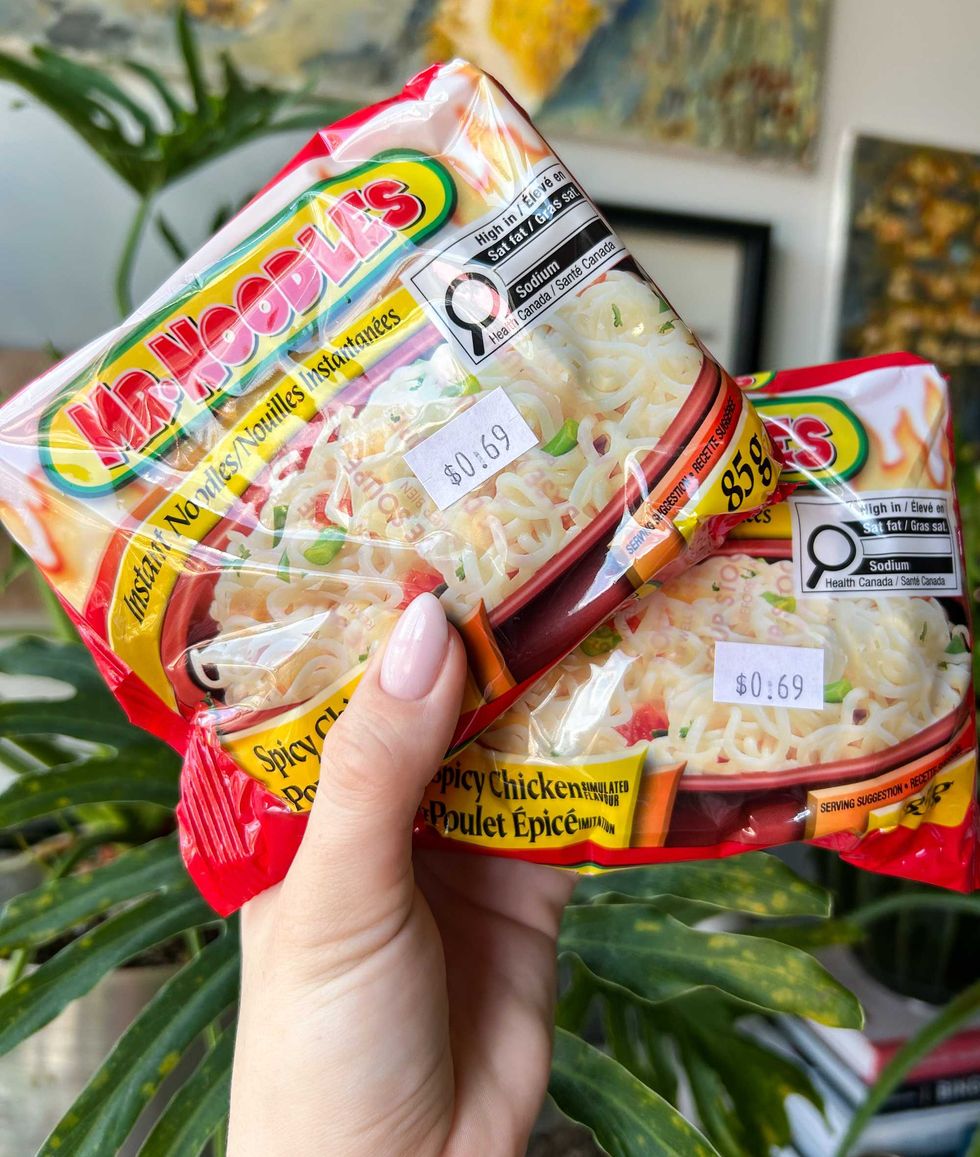 A photo of a person holding up two packets of Mr. Noodle Spicy Chicken instant noodles
