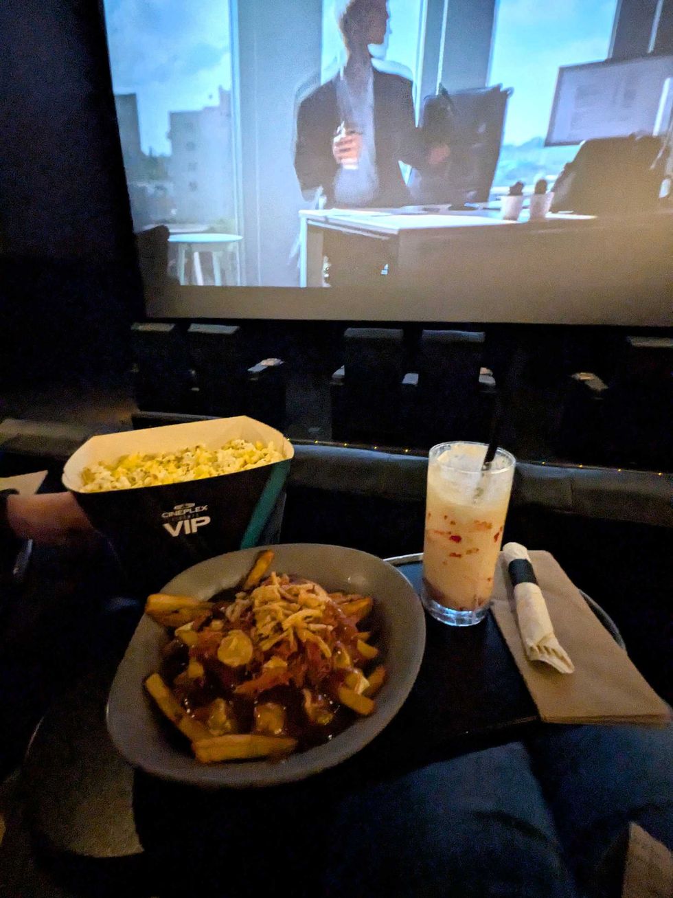 A plate of poutine, a milkshake, and box of popcorn on a table in front of a movie theatre screen