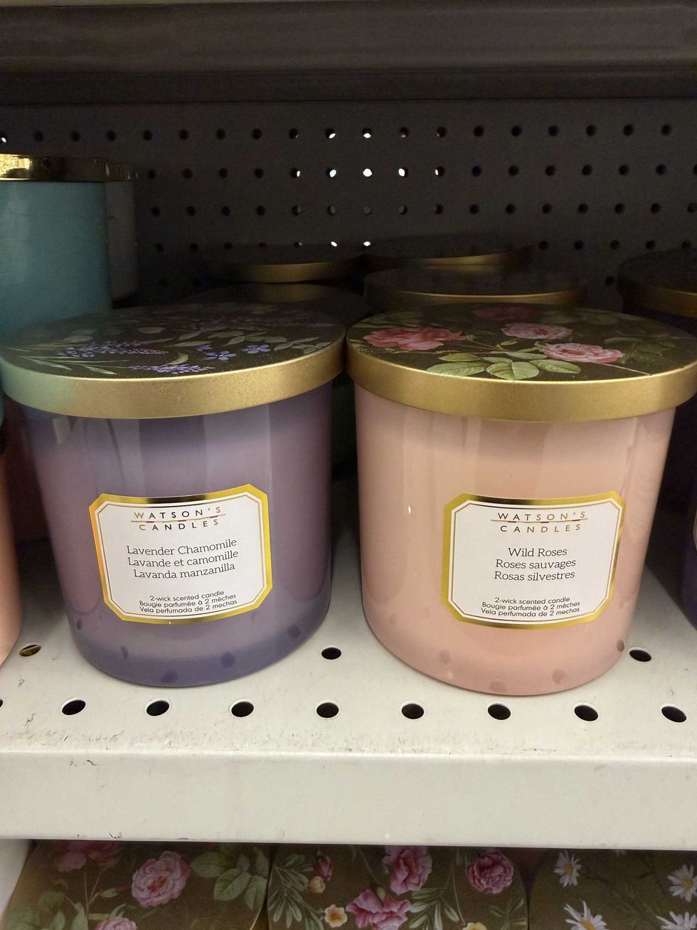 A purple candle next to a pink one with lids that have floral designs on them and one is lavender chamomile scented while the second is wild roses scented.