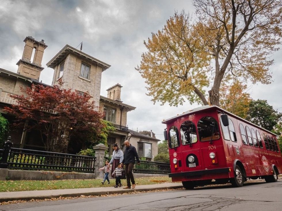 A red bus styled like a trolley car is pulled over on the street outside an old building. A young family walk along the pavement nearby. 