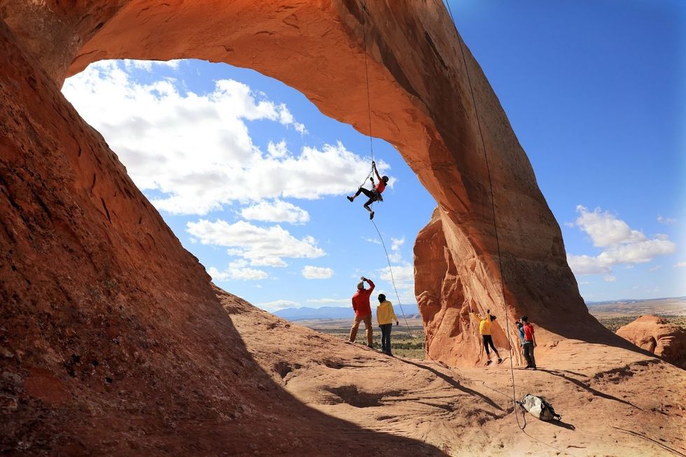 A rock climber climbs a rope underneath a natural sandstone arch in Arches National Park, USA.