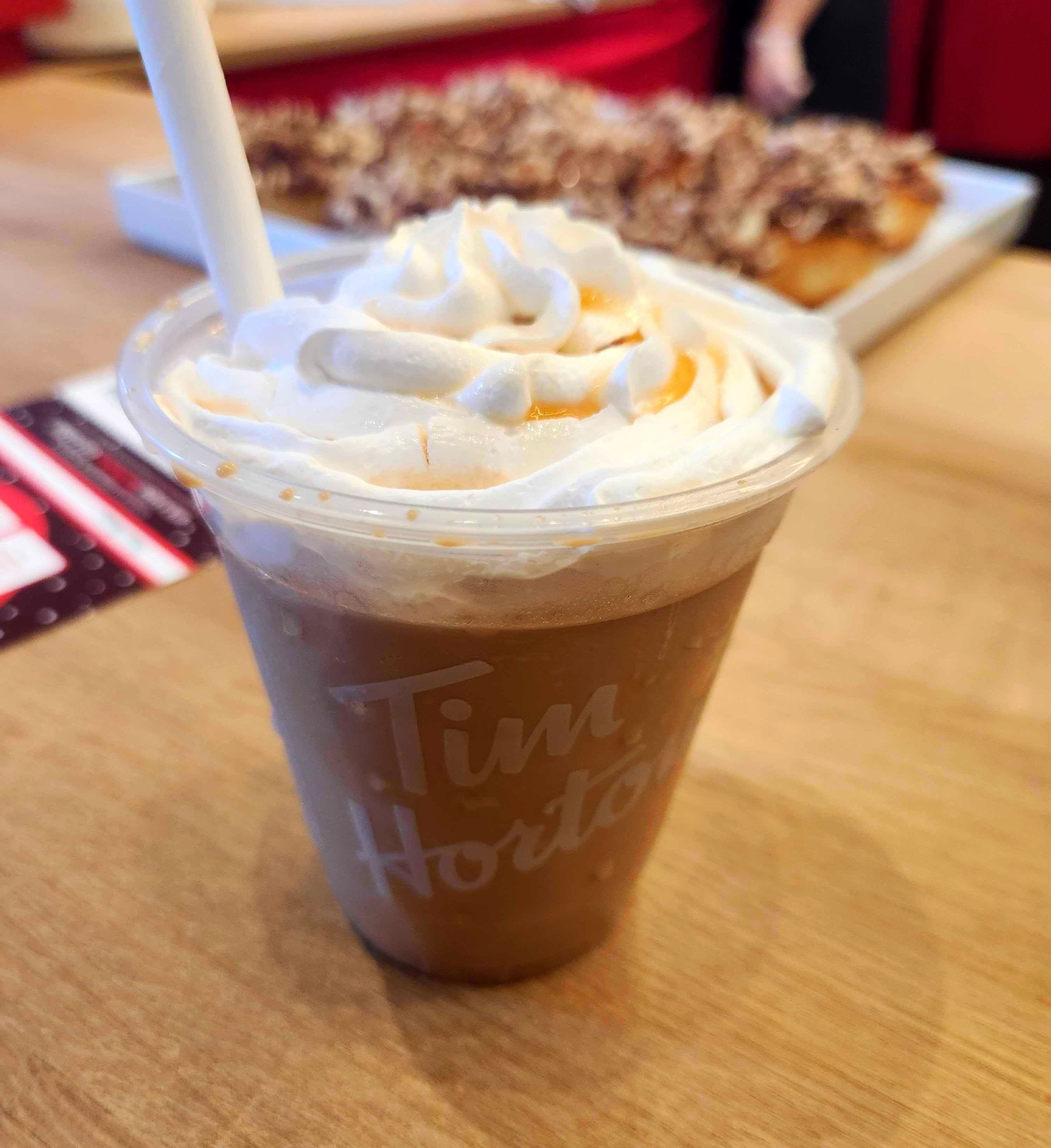 A see through coffee cup that has light brown iced capp in it with whipped cream on top and light brown syrup.