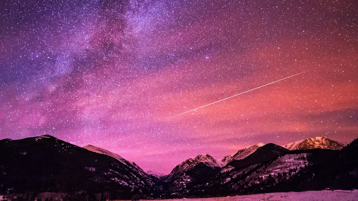 A shooting star is seen in a pink sky among tons of stars, with mountains in the background.