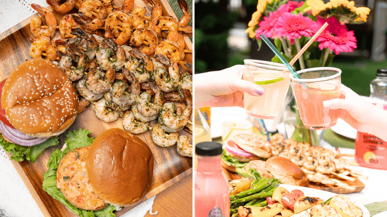 A summer barbecue spread featuring grilled shrimp, burgers, vegetables and two people toasting with colourful drinks outdoors.