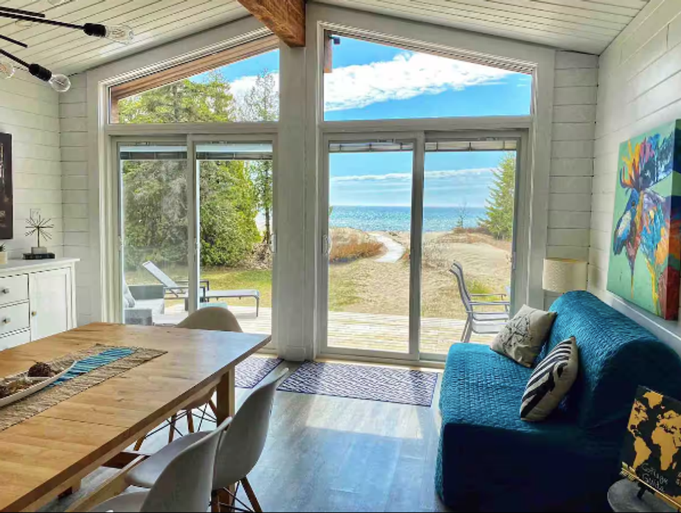 A table and sofa are seen in a cottage by a window with a view of a lake.