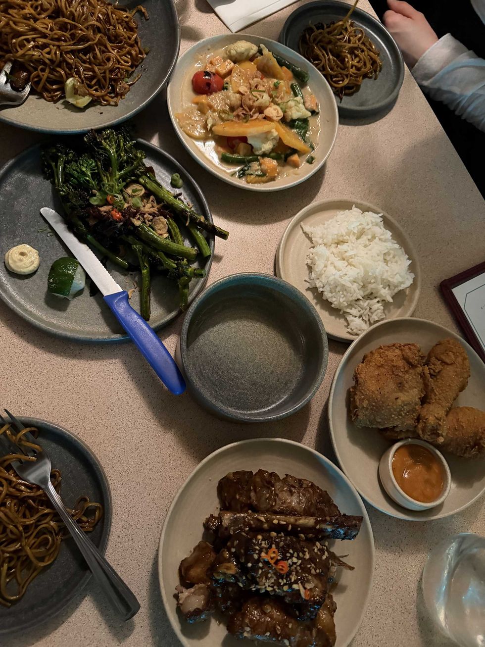 A tabletop filled with shareable dishes: fried chicken, pork ribs, noodles, broccolini, white steamed rice, and curry.