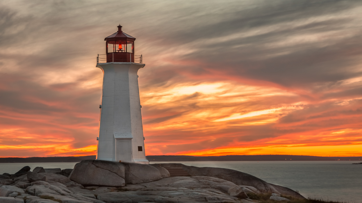 A tall white lighthouse stands on rocky shores, illuminated against a vibrant sunset sky with orange, red, and purple clouds reflecting over the ocean.