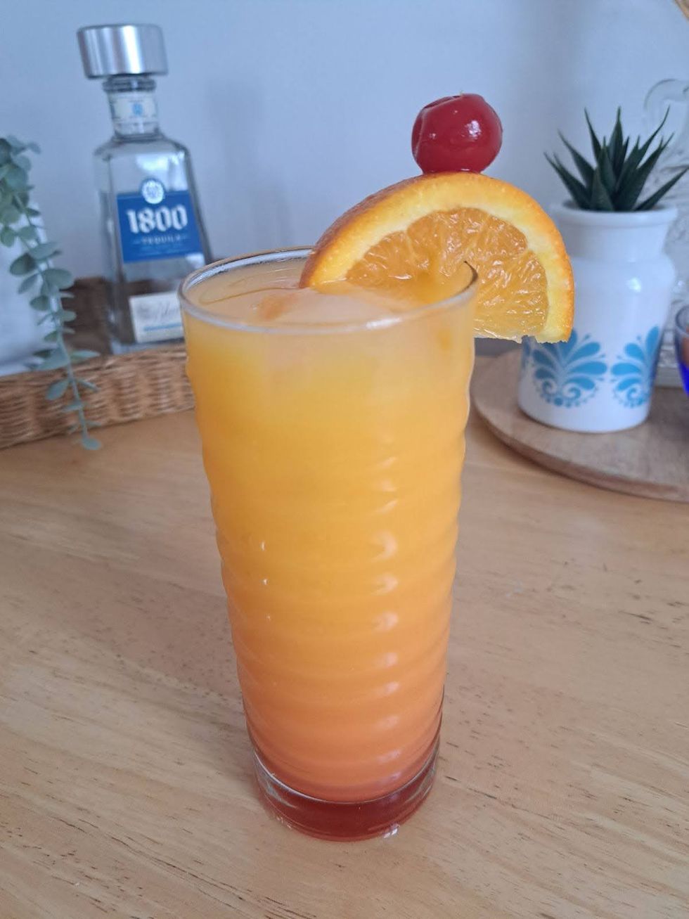 A tequila sunrise in a highball glass, garnished with a slice of orange and a cherry.
