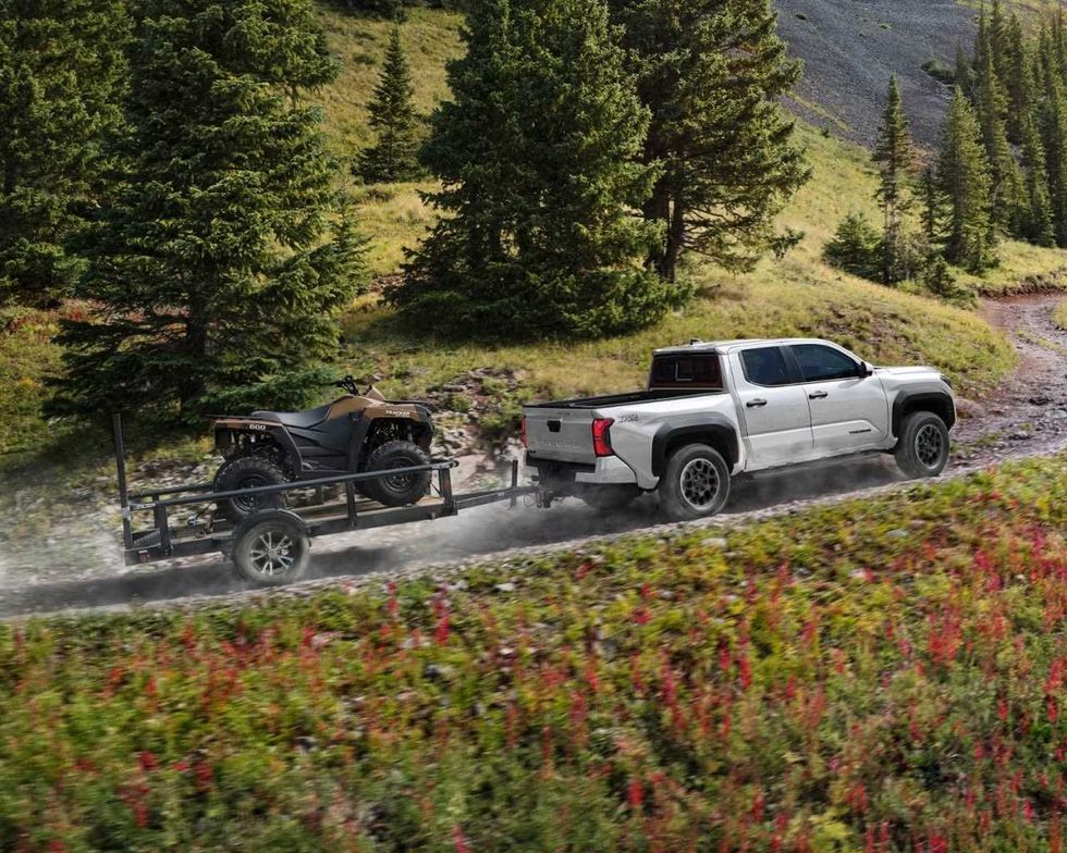 A Toyota Tacoma tows a flatbed trailer carrying a quad bike up a winding gravel hill.
