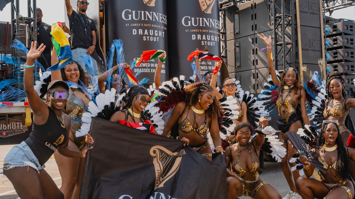 A vibrant group of costumed dancers posing in front of large Guinness Draught Stout displays at an outdoor event, wearing feathered headdresses, gold and black outfits, and smiling while holding a Guinness flag.