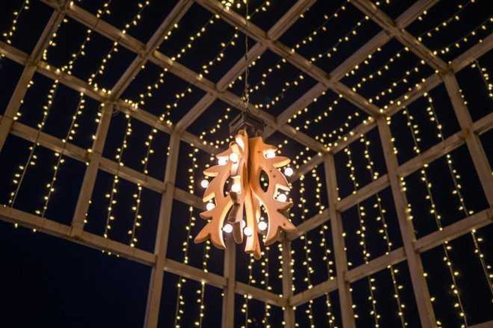 A warm wooden chandelier hangs from a grid-like ceiling draped with cascading strands of small golden fairy lights against a dark night background.