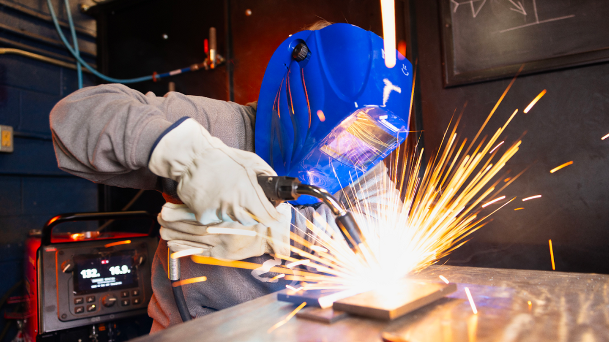 A welding student in a grey sweatshirt and blue welding mask uses a blow torch on a piece of metal.