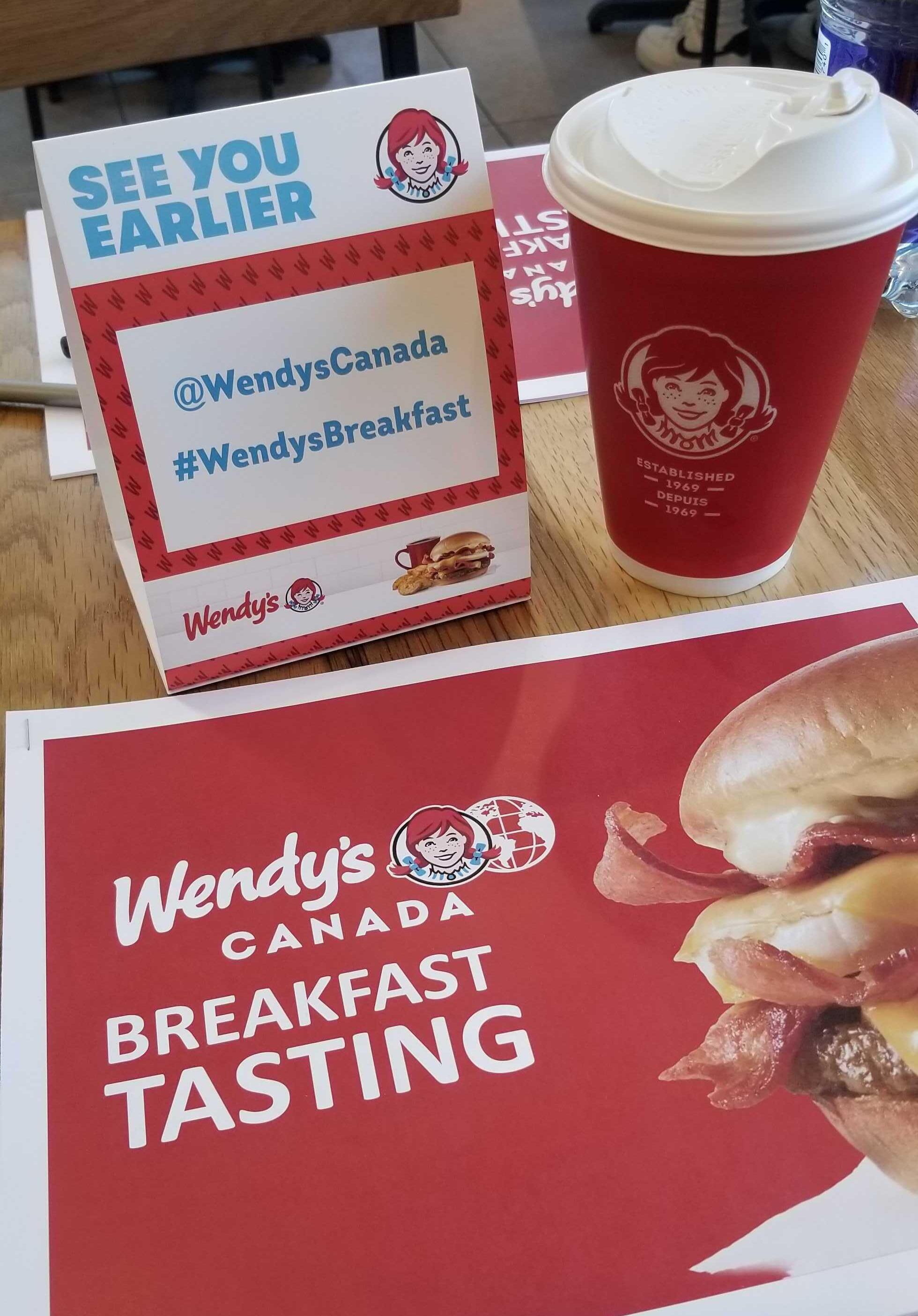 A Wendy's sign that says "see you earlier," a coffee, and a Wendy's Canada breakfast tasting package.