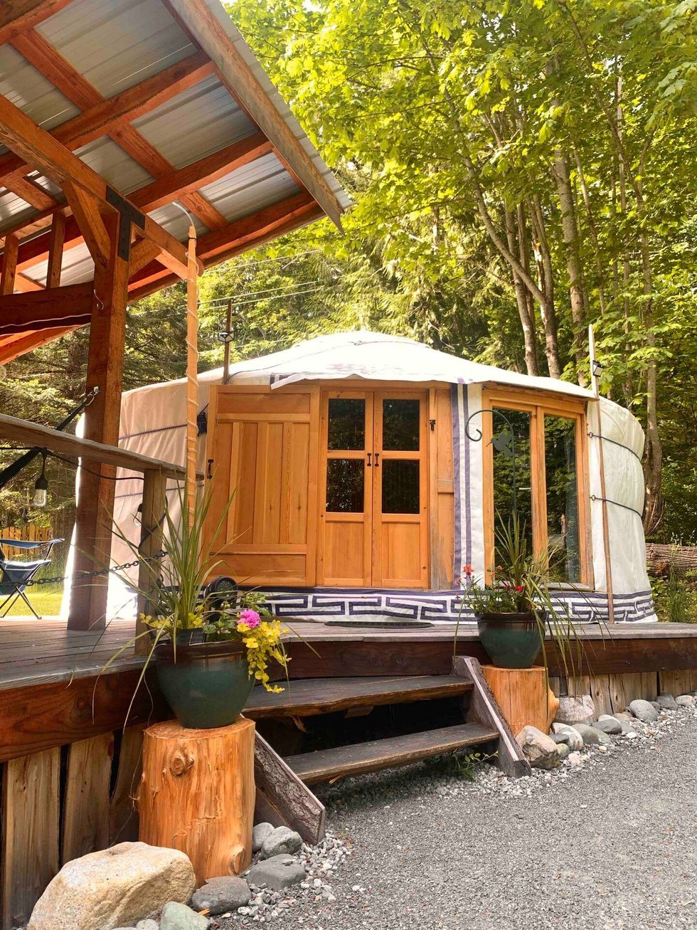 A white and brown yurt structure on a porch with trees and flower pots surrounding it.