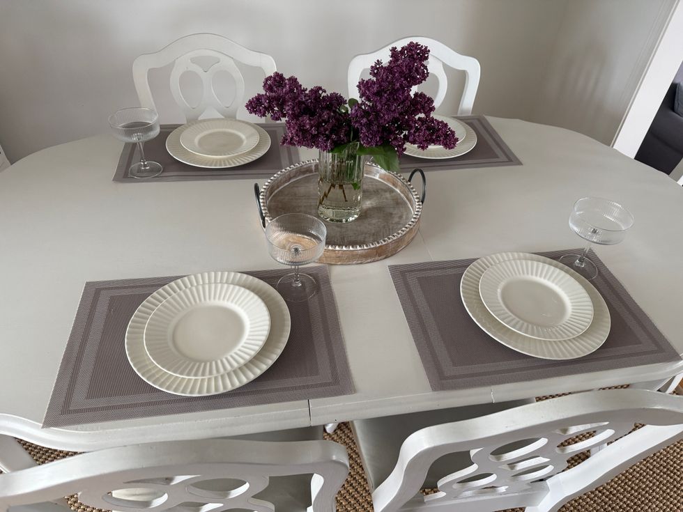A white dining table with grey placemats, white dishs and clear glasses with a tray and purple flowers in the centre.