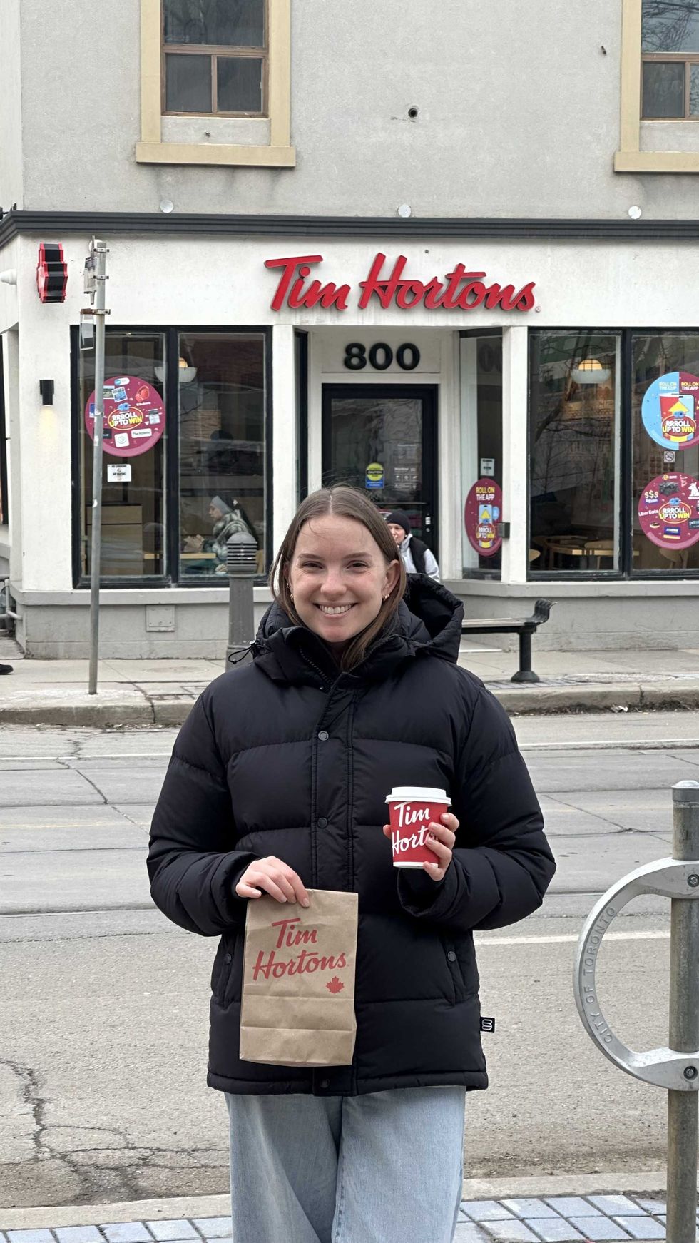 A woman holding a Tim Hortons bag and coffee cup while standing outside a Tim Hortons store.