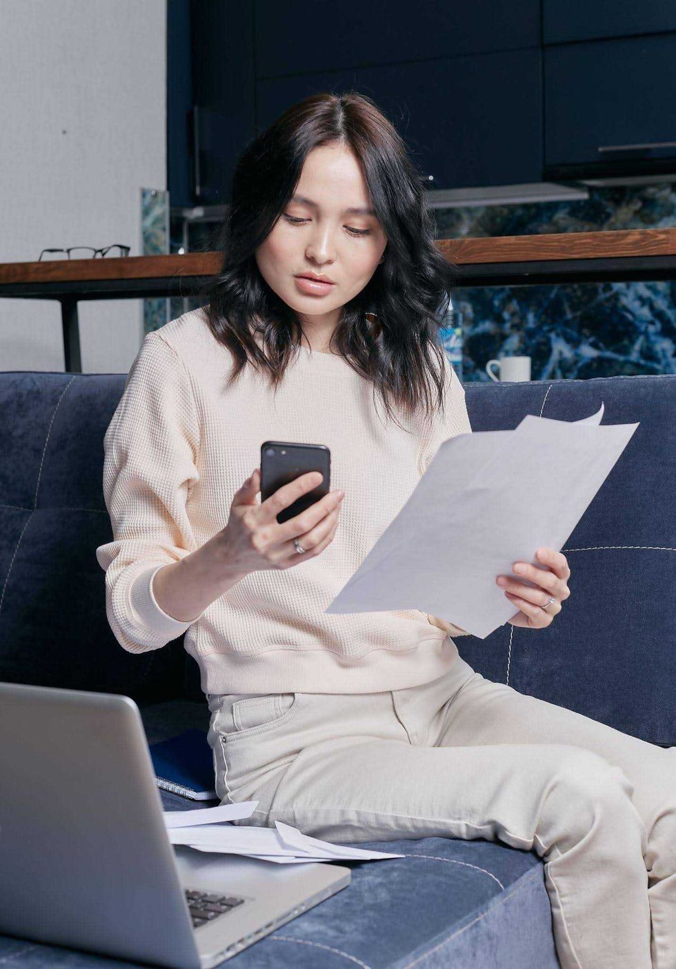 A woman holding papers looks at her phone next to an open laptop.