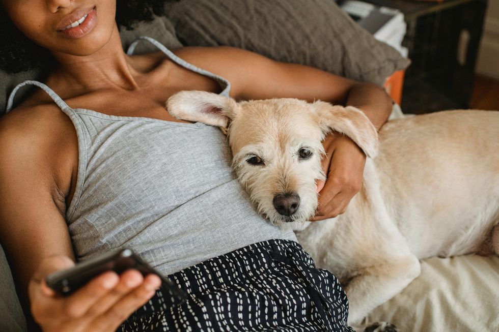 A woman in a grey singlet cuddles a white dog on a couch while looking at her phone.