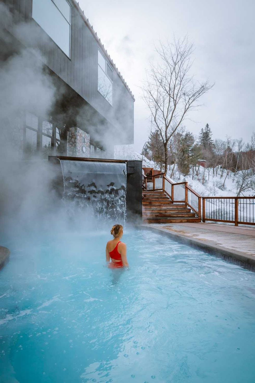 A woman in a red bathing suit in a hot pool at a Nordic spa surrounded by snow.