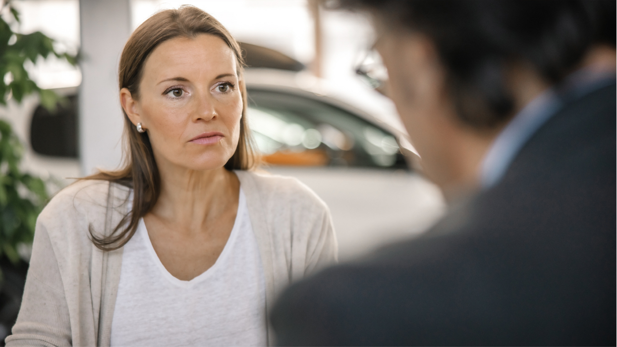 A woman in a white shirt and cardigan in a car show room with a serious look on her face.