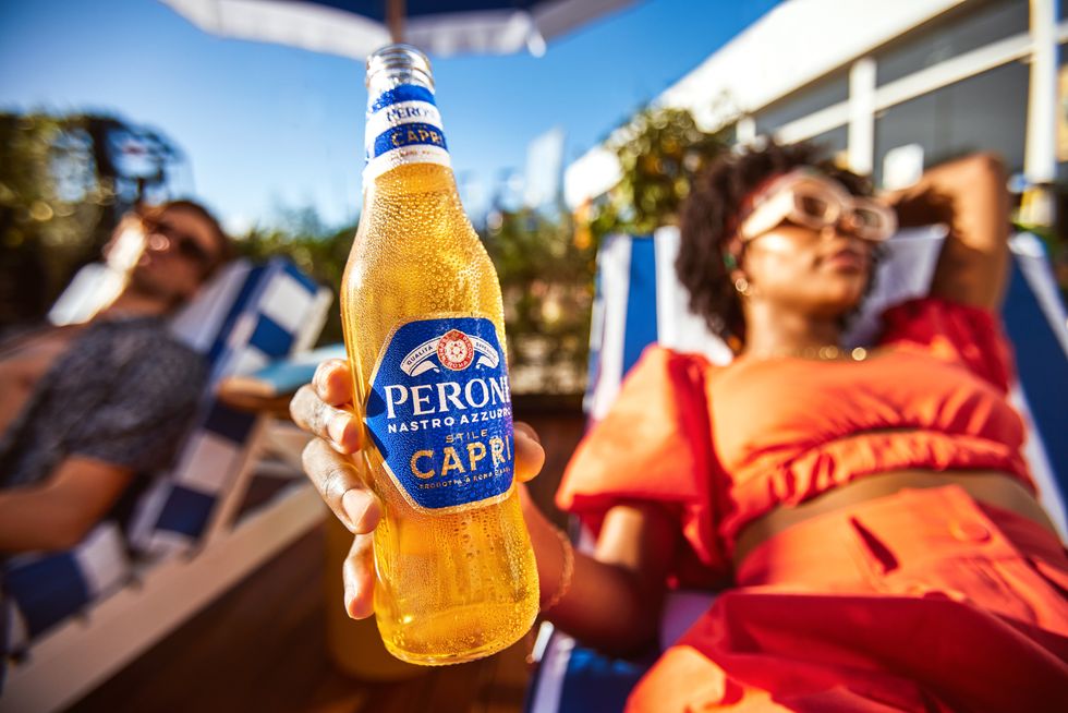 A woman in an orange dress reclines on a white and blue striped sun lounger, holding a bottle of Peroni Nasto Azzurro Stile Capri.
