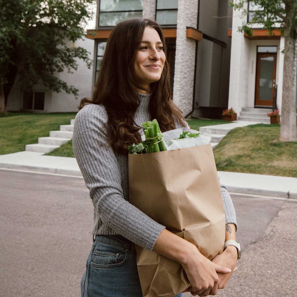 A woman in jeans and a sweater stands on a street with a bag of groceries in her arms.