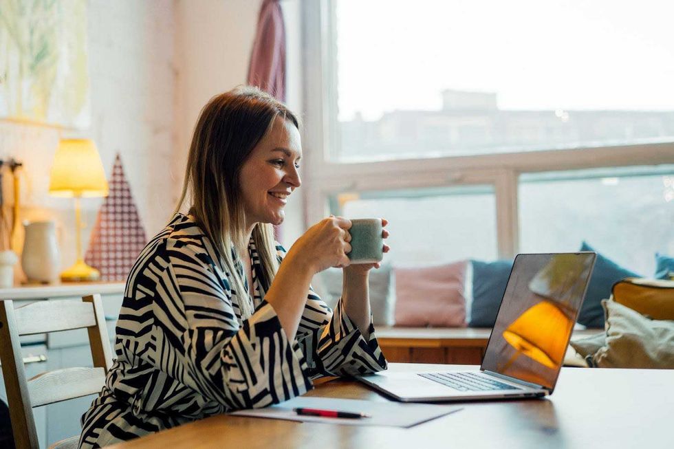 A woman sits at a dining table with a cup of tea, speaking to someone on a video chat.