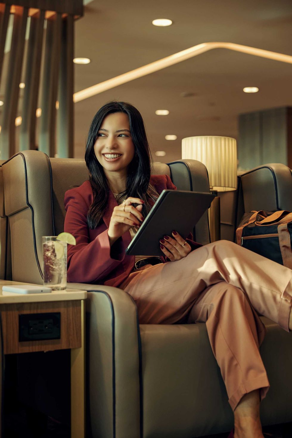 A woman sits in a leather chair with a tablet and drink.
