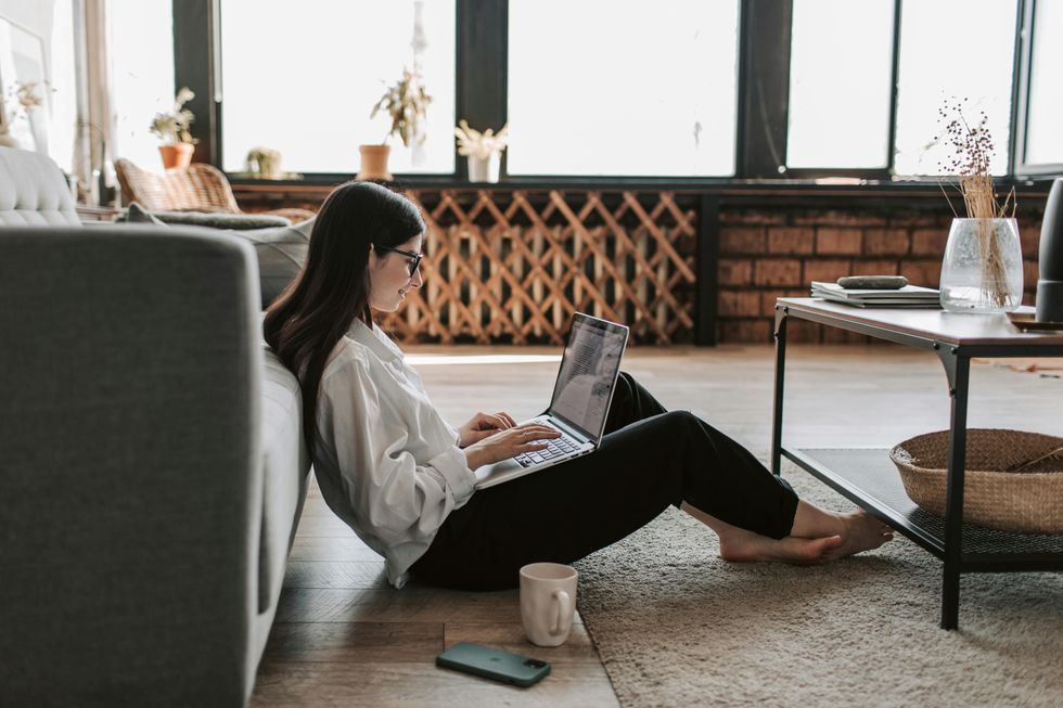 A woman sits on the floor in front of a couch doing work on a laptop.