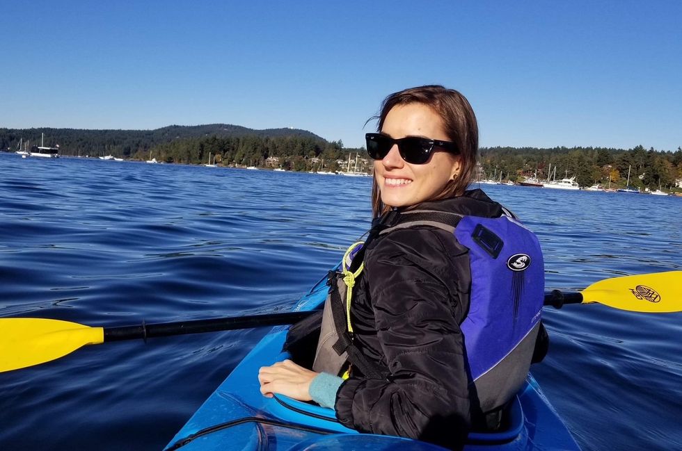 A woman sitting in a blue kayak on the blue water with a blue life jacket and black and yellow paddle.